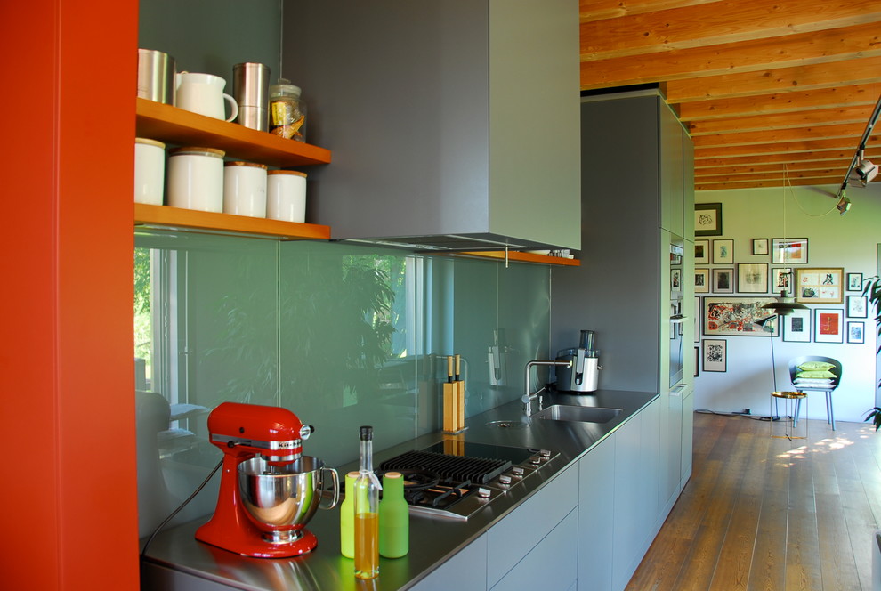 Foto på ett mellanstort funkis linjärt kök med öppen planlösning, med en integrerad diskho, släta luckor, grå skåp, bänkskiva i rostfritt stål, grönt stänkskydd, glaspanel som stänkskydd och mörkt trägolv