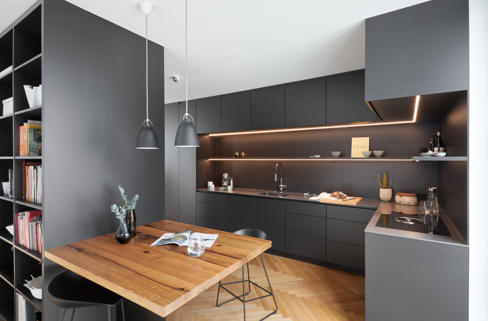 На фото: угловая кухня-гостиная в современном стиле с монолитной мойкой, плоскими фасадами, черными фасадами, столешницей из нержавеющей стали, черным фартуком и светлым паркетным полом