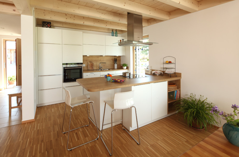 Inspiration för moderna linjära brunt kök med öppen planlösning, med släta luckor, vita skåp, träbänkskiva, vitt stänkskydd, stänkskydd i trä, ljust trägolv, en nedsänkt diskho, svarta vitvaror, en halv köksö och brunt golv
