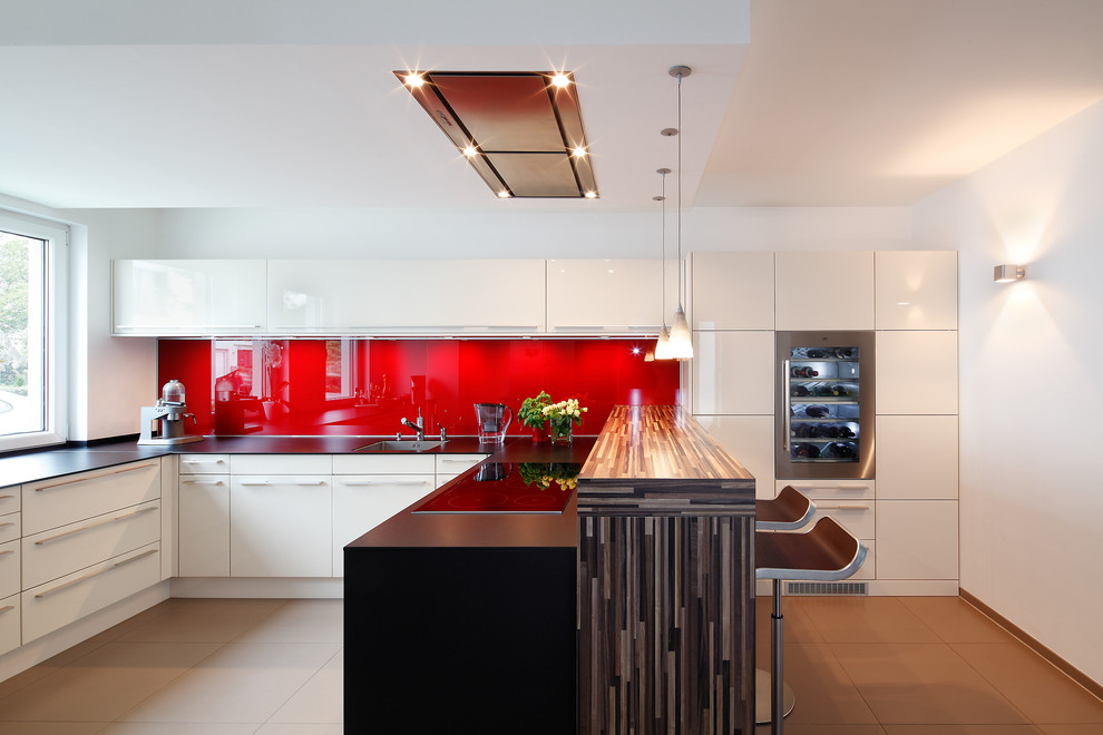 Large contemporary l-shaped enclosed kitchen in Dusseldorf with a submerged sink, flat-panel cabinets, white cabinets, red splashback, glass sheet splashback, integrated appliances, a breakfast bar and beige floors.