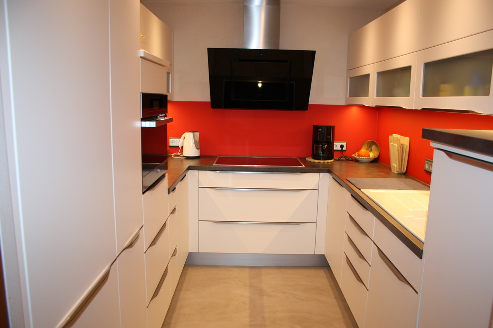 Foto på ett avskilt funkis u-kök, med vita skåp, träbänkskiva, rött stänkskydd, glaspanel som stänkskydd och rostfria vitvaror