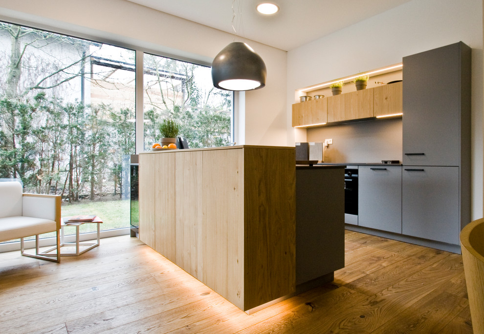 Cette image montre une cuisine américaine parallèle design en bois clair avec un placard à porte plane, une crédence grise, un électroménager noir, un sol en bois brun, une péninsule, un sol marron et plan de travail noir.