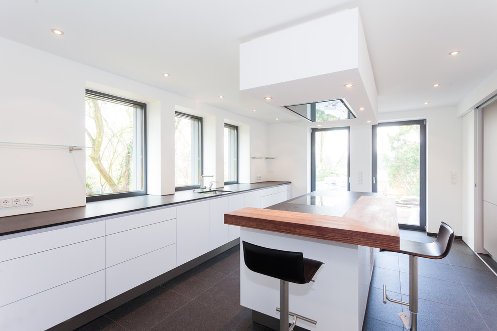 На фото: параллельная кухня-гостиная в современном стиле с белыми фасадами, деревянной столешницей, белым фартуком, островом и черной техникой