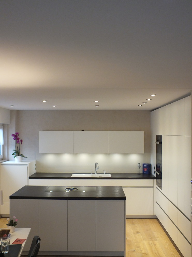 Inspiration for a contemporary galley open plan kitchen in Dortmund with a built-in sink, white cabinets, beige splashback, glass sheet splashback, light hardwood flooring and an island.