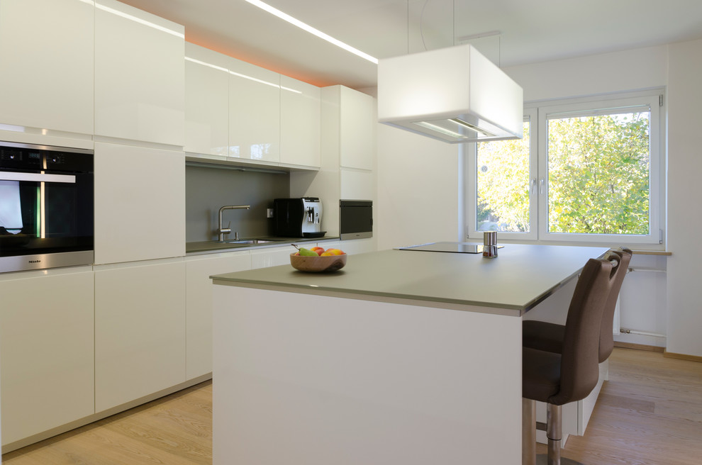 Inspiration for a medium sized contemporary single-wall kitchen/diner in Nuremberg with flat-panel cabinets, white cabinets, grey splashback, stainless steel appliances, light hardwood flooring, an island and a submerged sink.