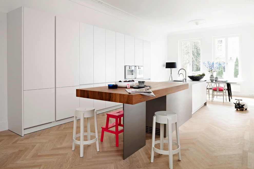 Große Moderne Wohnküche mit flächenbündigen Schrankfronten, weißen Schränken, hellem Holzboden, Kücheninsel, Unterbauwaschbecken und Elektrogeräten mit Frontblende in Hamburg