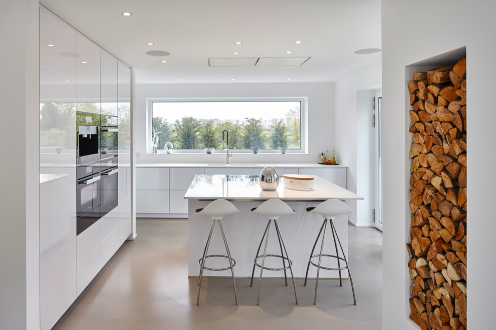 Свежая идея для дизайна: угловая кухня-гостиная среднего размера в современном стиле с плоскими фасадами, белыми фасадами, белым фартуком, черной техникой, бетонным полом и островом - отличное фото интерьера