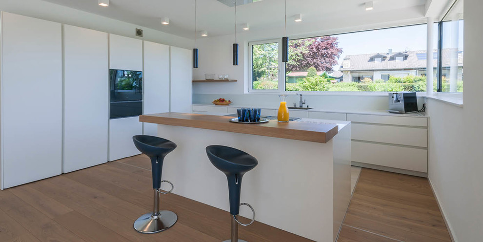 This is an example of a medium sized contemporary kitchen/diner in Other with flat-panel cabinets, white cabinets, medium hardwood flooring, an island, a built-in sink, wood worktops, white splashback and black appliances.