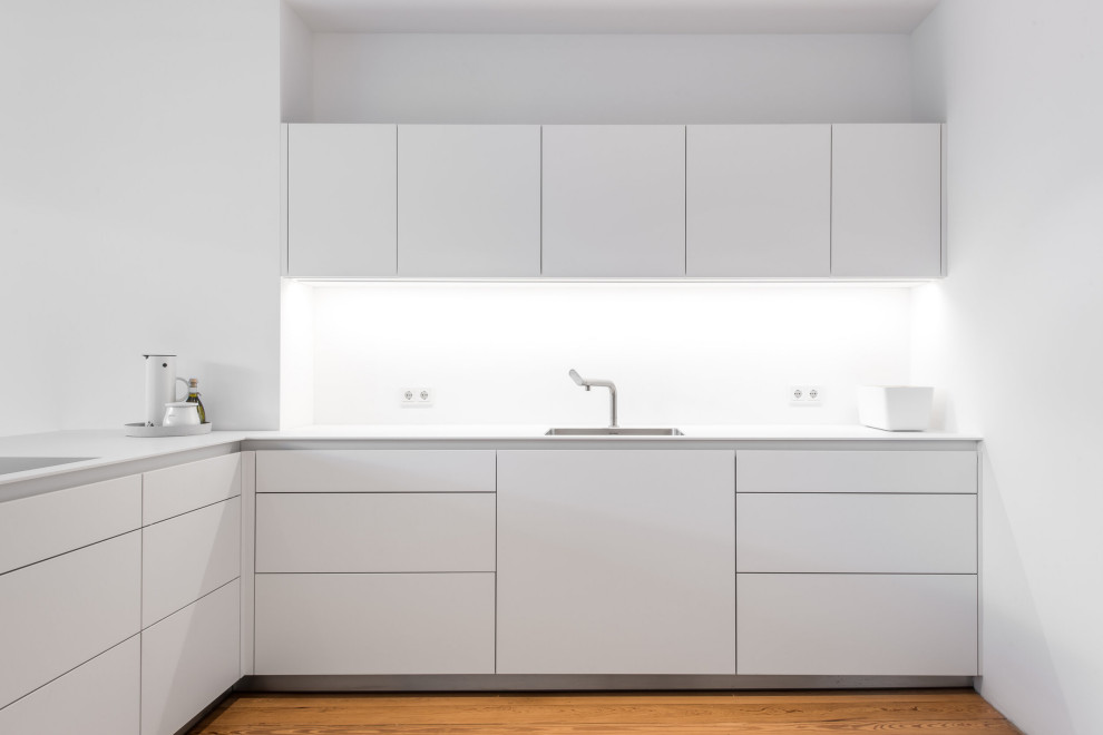 Mid-sized minimalist l-shaped light wood floor open concept kitchen photo in Hamburg with a single-bowl sink, flat-panel cabinets, white cabinets, solid surface countertops, white backsplash, glass sheet backsplash, white appliances, a peninsula and white countertops