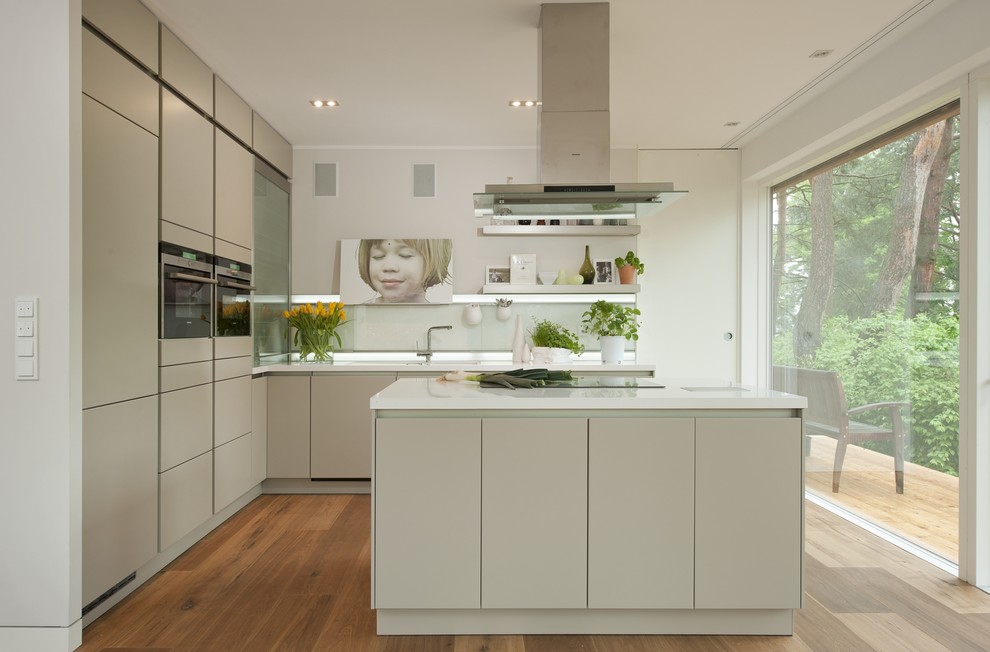 Inredning av ett modernt stort kök med öppen planlösning, med släta luckor, grå skåp, vitt stänkskydd, glaspanel som stänkskydd, rostfria vitvaror, mellanmörkt trägolv och en köksö