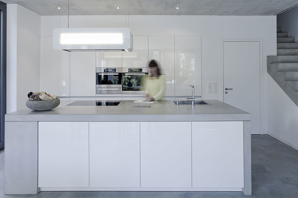 Идея дизайна: кухня-гостиная среднего размера в стиле модернизм с накладной мойкой, плоскими фасадами, белыми фасадами, столешницей из бетона, техникой под мебельный фасад, бетонным полом, островом и серым полом