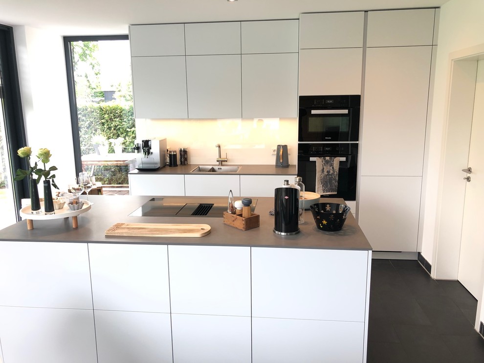 Idée de décoration pour une cuisine américaine parallèle minimaliste de taille moyenne avec un évier intégré, un placard à porte plane, des portes de placard blanches, une crédence blanche, une crédence en feuille de verre, un électroménager noir, un sol en carrelage de porcelaine, îlot, un sol gris et un plan de travail gris.