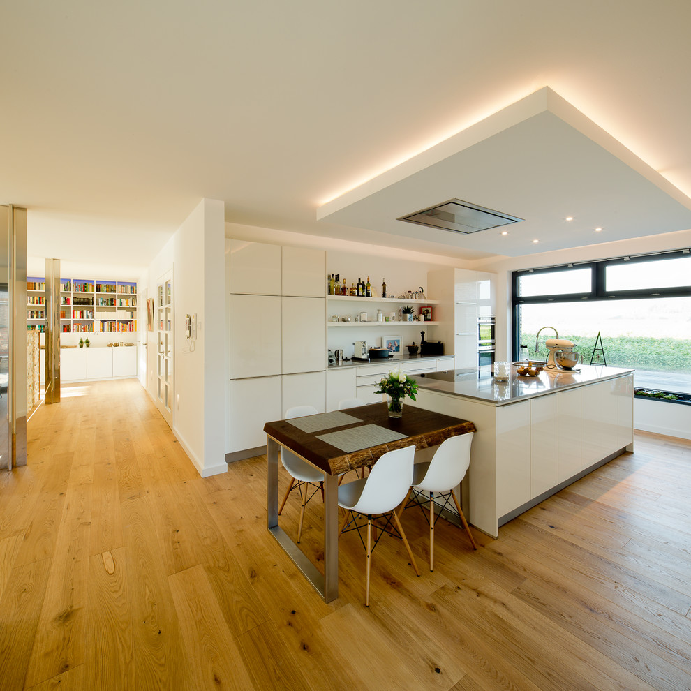 This is an example of a contemporary galley open plan kitchen in Dusseldorf with flat-panel cabinets, white cabinets, white splashback, integrated appliances, light hardwood flooring and an island.