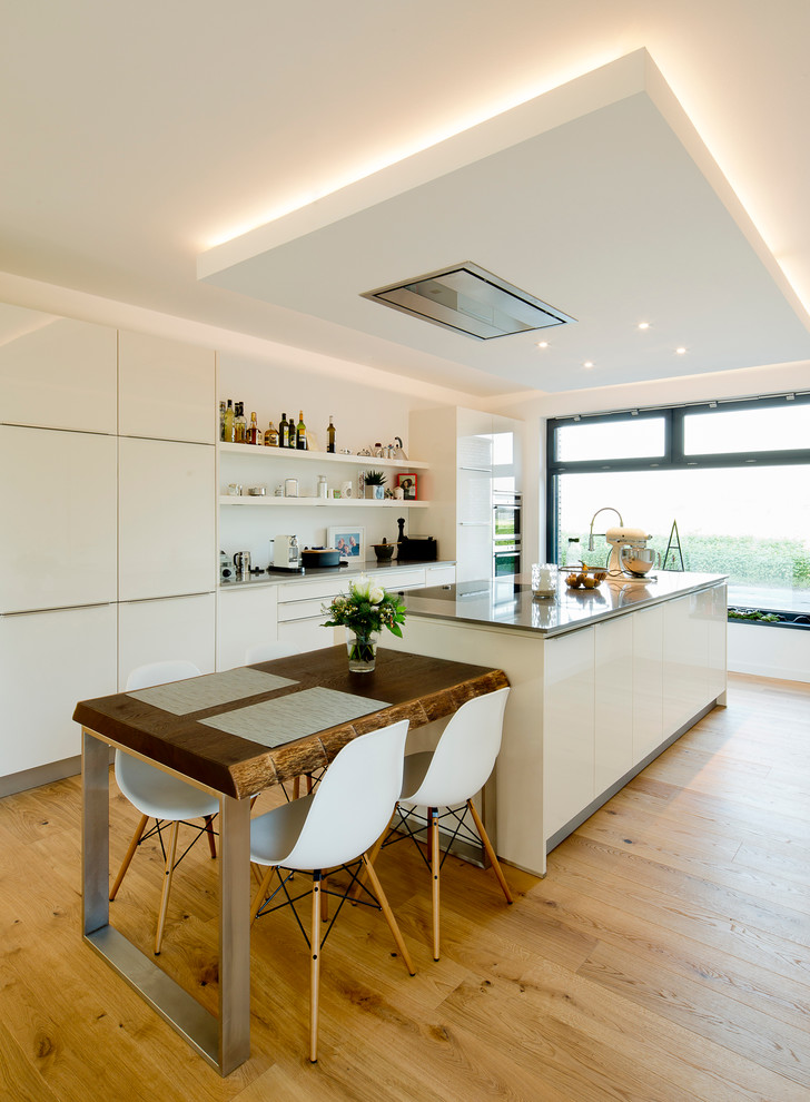 Photo of a large contemporary single-wall kitchen/diner in Dusseldorf with medium hardwood flooring, an island, flat-panel cabinets, white cabinets, white splashback and stainless steel appliances.