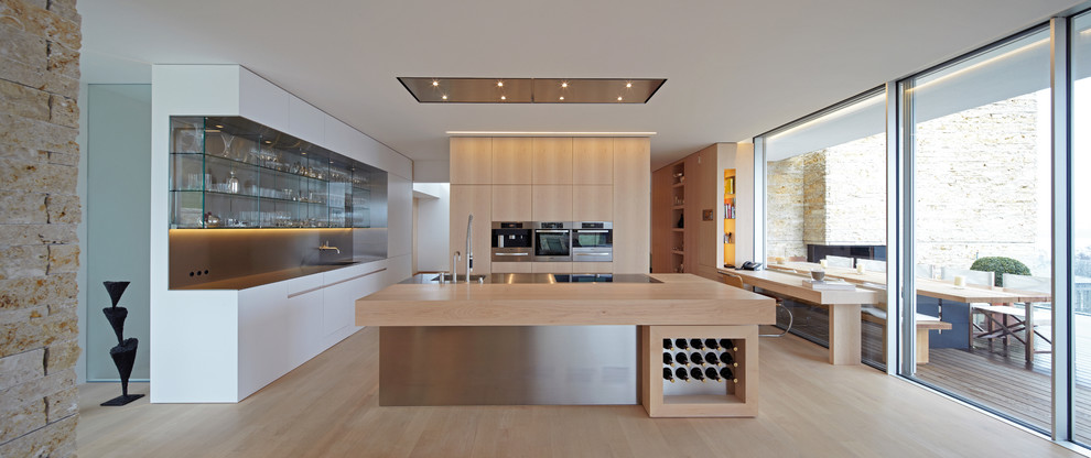 Foto på ett mycket stort minimalistiskt kök med öppen planlösning, med en integrerad diskho, släta luckor, skåp i mellenmörkt trä, bänkskiva i rostfritt stål, ljust trägolv och en köksö