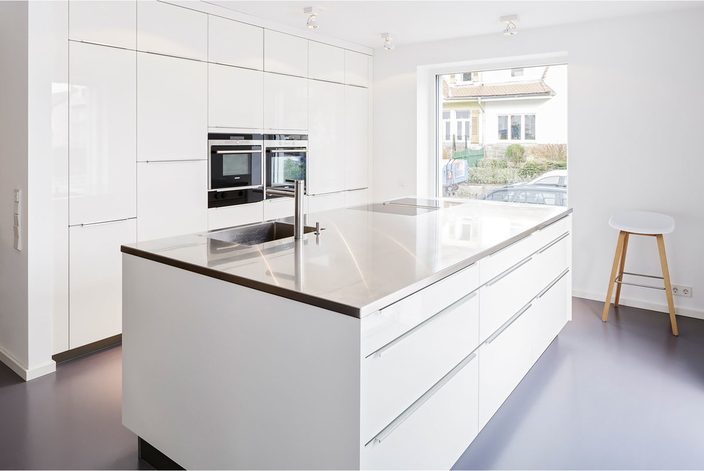 Large contemporary single-wall open plan kitchen in Other with an integrated sink, flat-panel cabinets, white cabinets, black appliances, an island and stainless steel worktops.