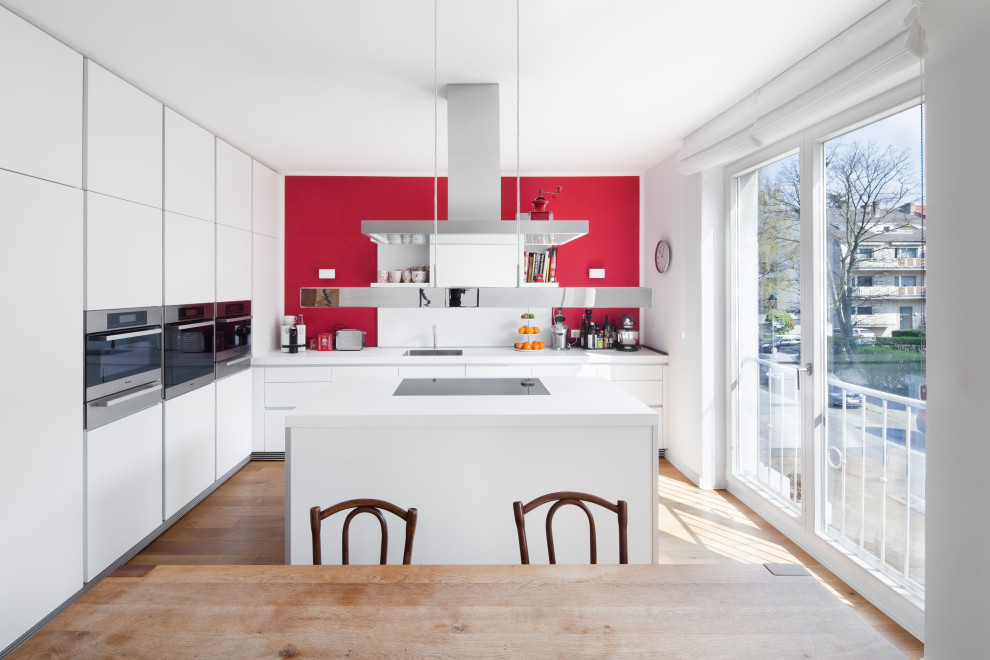 Moderne Wohnküche mit flächenbündigen Schrankfronten, weißen Schränken, Küchengeräten aus Edelstahl, Kücheninsel, Waschbecken, Küchenrückwand in Rot, hellem Holzboden und weißer Arbeitsplatte in Düsseldorf