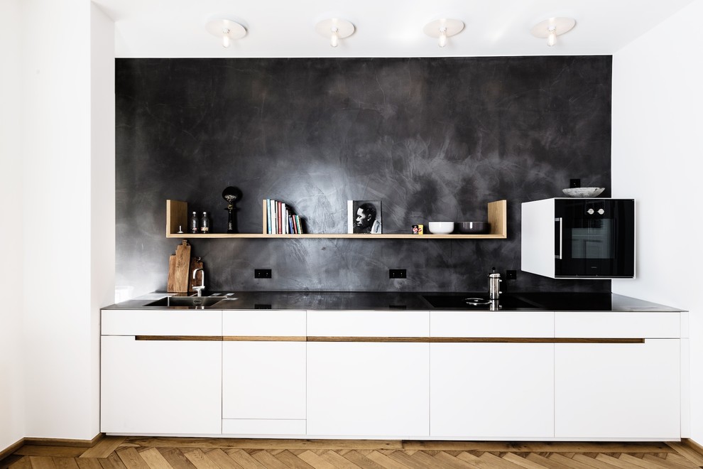 Exempel på ett litet modernt linjärt kök med öppen planlösning, med en integrerad diskho, släta luckor, vita skåp, bänkskiva i rostfritt stål, svart stänkskydd, stänkskydd i kalk, svarta vitvaror, mellanmörkt trägolv och brunt golv
