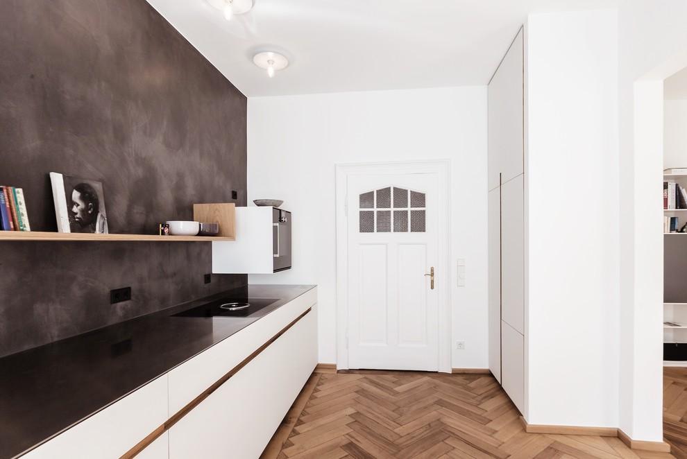 Photo of a large modern single-wall open plan kitchen in Munich with an integrated sink, flat-panel cabinets, white cabinets, stainless steel worktops, black splashback, limestone splashback, black appliances, medium hardwood flooring, no island and brown floors.