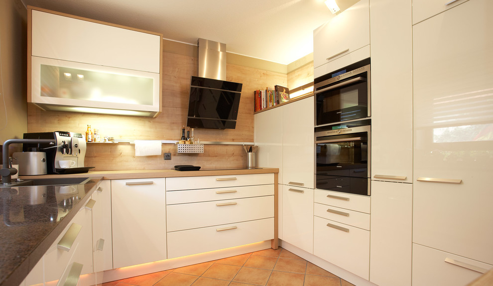 ニュルンベルクにあるコンテンポラリースタイルのおしゃれなキッチン (ドロップインシンク、白いキャビネット、茶色いキッチンパネル、木材のキッチンパネル、黒い調理設備、テラコッタタイルの床、アイランドなし、ベージュの床) の写真