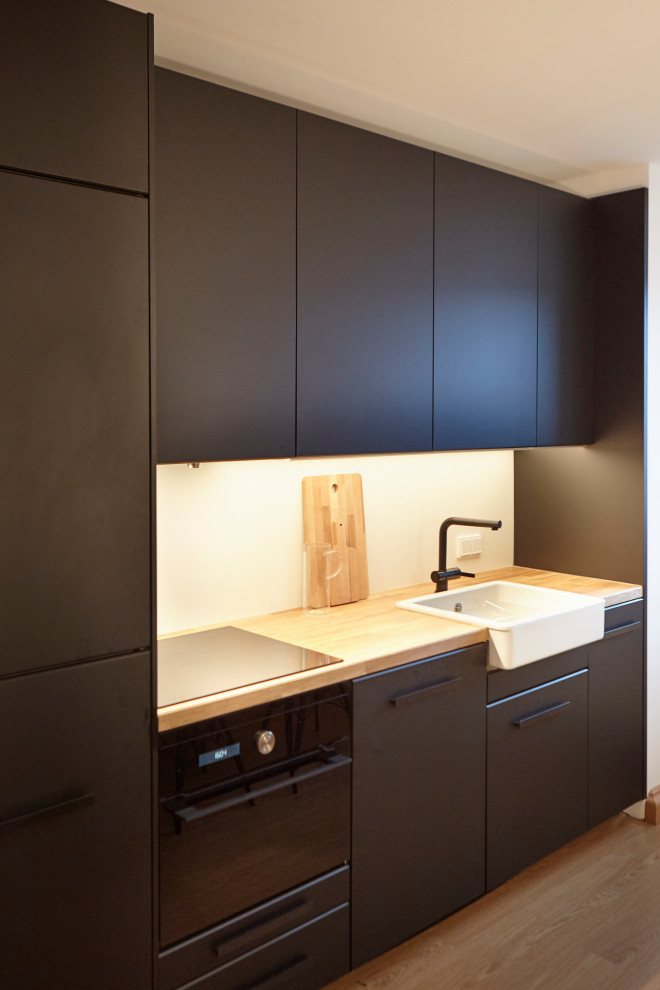 ミュンヘンにある高級な小さな北欧スタイルのおしゃれなキッチン (エプロンフロントシンク、フラットパネル扉のキャビネット、黒いキャビネット、木材カウンター、白いキッチンパネル、ガラス板のキッチンパネル、黒い調理設備、無垢フローリング、アイランドなし、ベージュの床、ベージュのキッチンカウンター) の写真