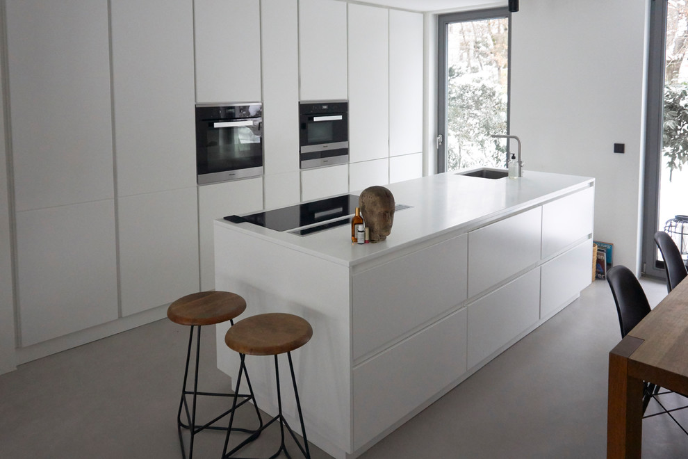 This is an example of an expansive modern galley open plan kitchen in Munich with a submerged sink, flat-panel cabinets, white cabinets, composite countertops, white splashback, stainless steel appliances, concrete flooring, an island, grey floors, white worktops and brick splashback.