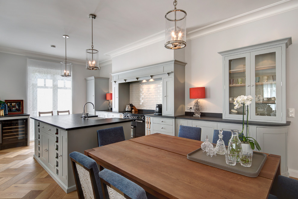 Photo of a large traditional single-wall kitchen/diner in Berlin with grey cabinets, white splashback, metro tiled splashback, light hardwood flooring, an island, a belfast sink, shaker cabinets, black appliances, wood worktops and brown floors.
