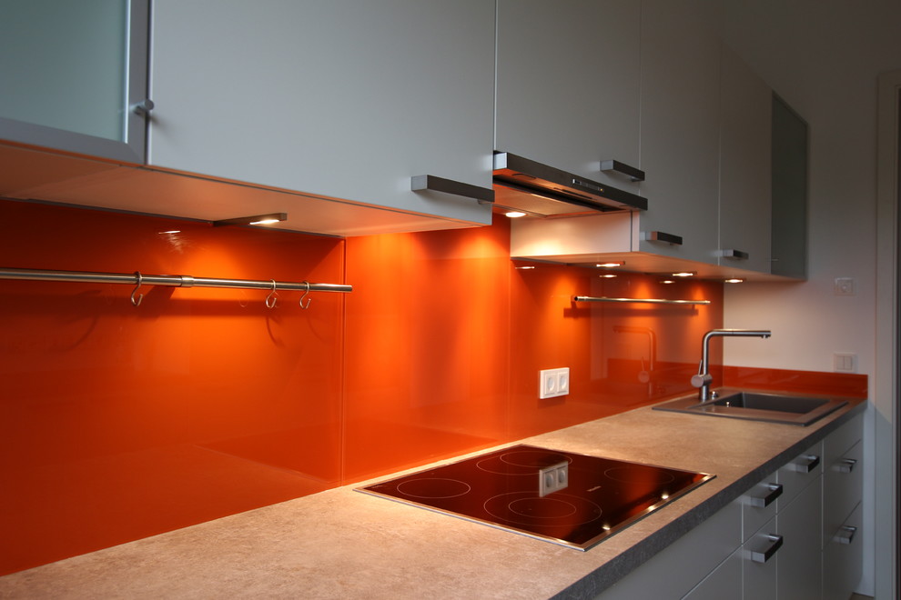 他の地域にある小さなコンテンポラリースタイルのおしゃれなII型キッチン (フラットパネル扉のキャビネット、ラミネートカウンター、オレンジのキッチンパネル、ガラス板のキッチンパネル、シルバーの調理設備) の写真