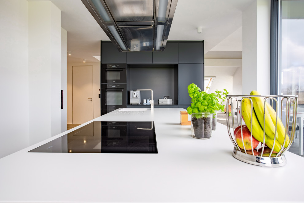 Large modern open plan kitchen in Dortmund with an integrated sink, flat-panel cabinets, white cabinets, composite countertops, black appliances, painted wood flooring, an island and white worktops.