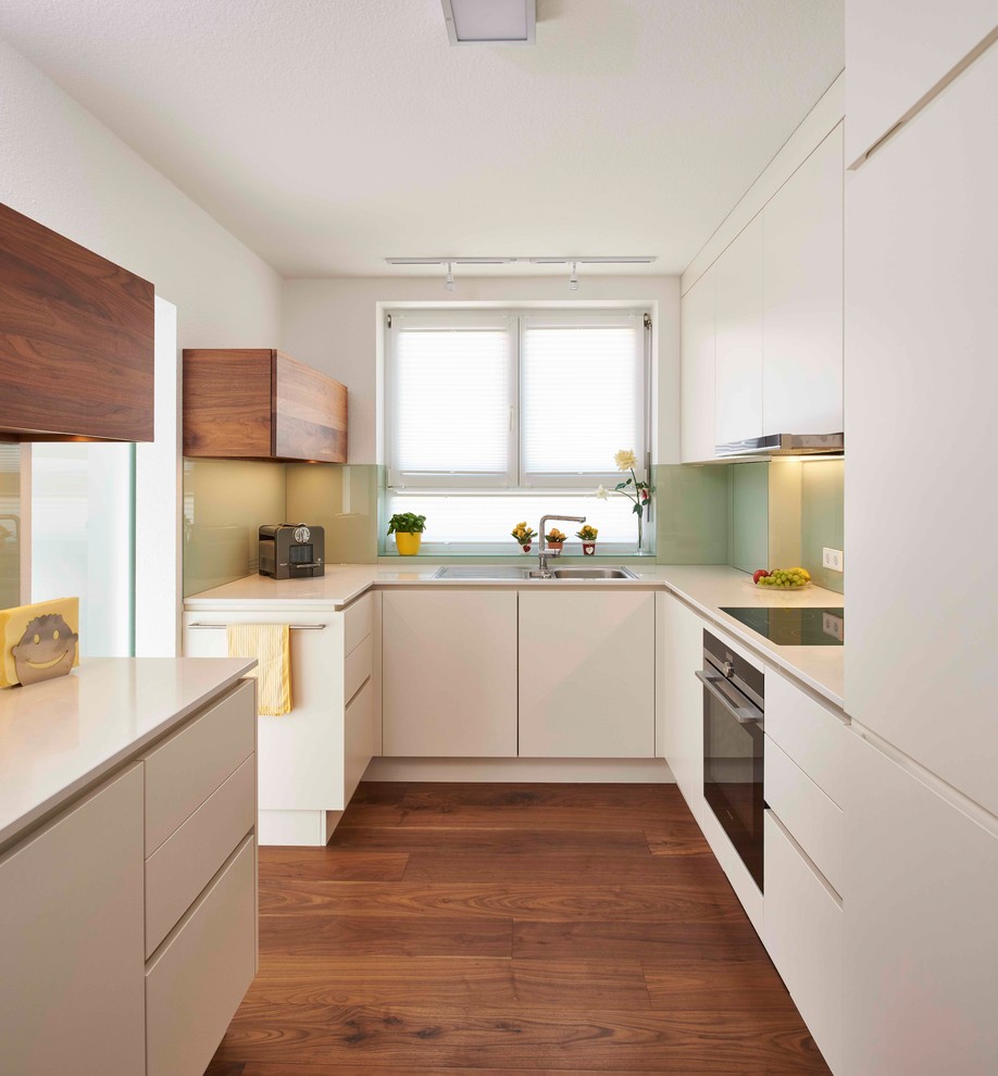 Design ideas for a small contemporary u-shaped enclosed kitchen in Stuttgart with a built-in sink, flat-panel cabinets, white cabinets, green splashback, glass sheet splashback, medium hardwood flooring, no island, brown floors, white worktops and integrated appliances.
