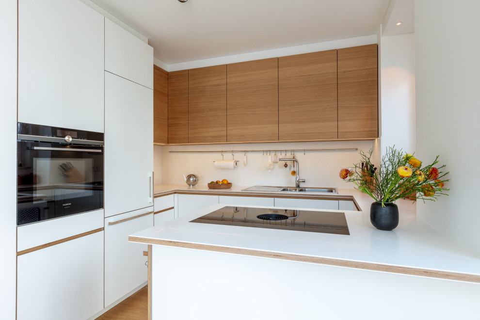 Offene, Mittelgroße Moderne Küche in U-Form mit Einbauwaschbecken, flächenbündigen Schrankfronten, weißen Schränken, Mineralwerkstoff-Arbeitsplatte, Küchenrückwand in Weiß, schwarzen Elektrogeräten, braunem Holzboden, Kücheninsel, braunem Boden, weißer Arbeitsplatte und eingelassener Decke in München