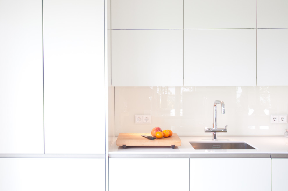 Diseño de cocina minimalista abierta sin isla con fregadero integrado, puertas de armario blancas, encimera de vidrio, salpicadero blanco, salpicadero de vidrio templado y encimeras blancas