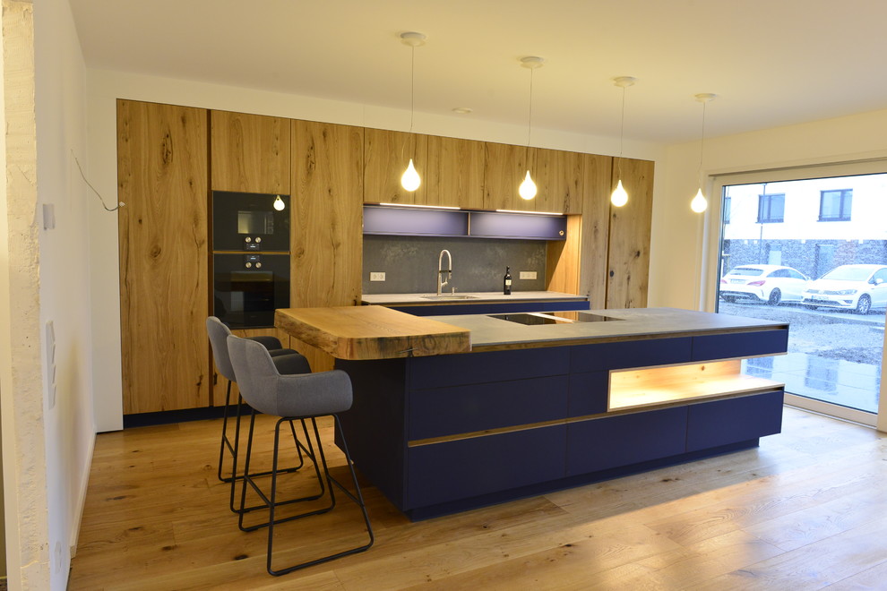 Expansive modern galley open plan kitchen in Dortmund with an integrated sink, flat-panel cabinets, blue cabinets, composite countertops, white splashback, black appliances, dark hardwood flooring, an island, brown floors and grey worktops.