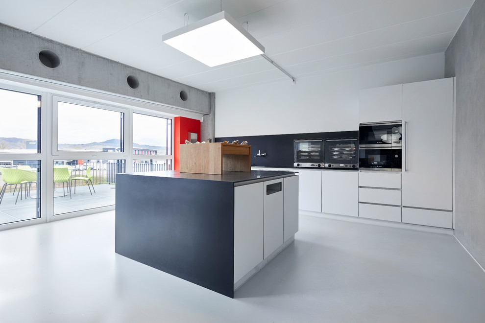 На фото: отдельная, прямая кухня среднего размера в стиле модернизм с плоскими фасадами, белыми фасадами, черным фартуком, черной техникой, бетонным полом, островом, серым полом и черной столешницей с