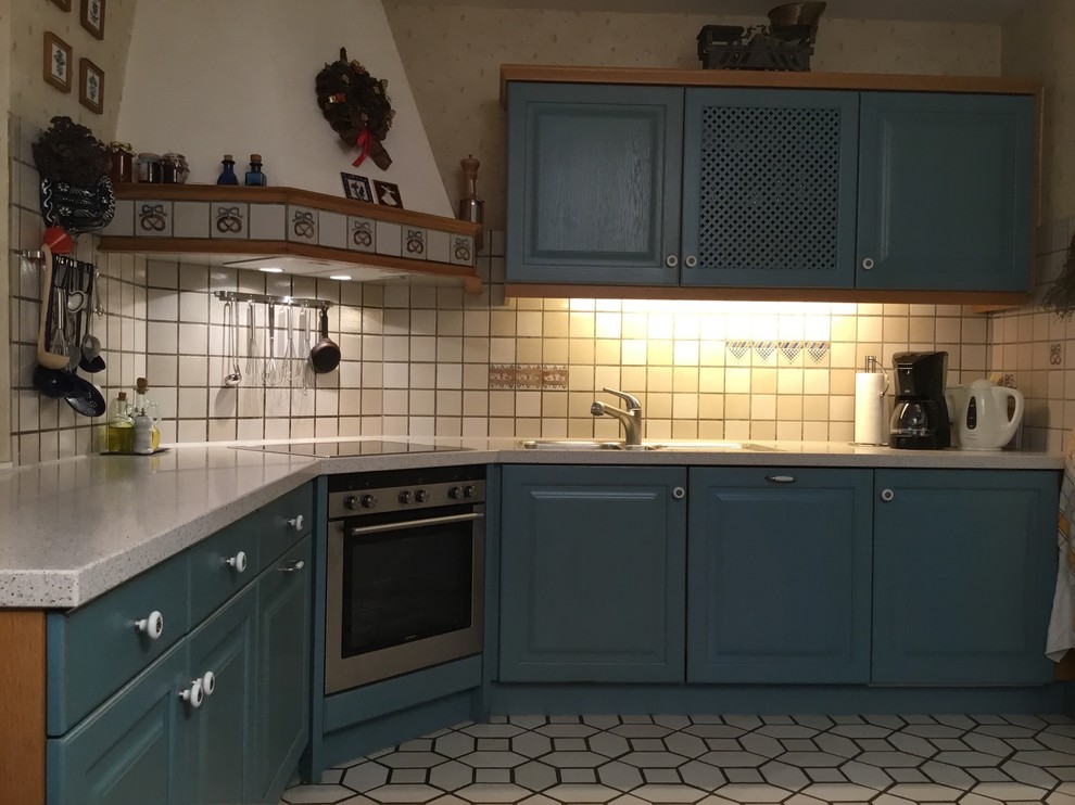 Photo of a medium sized farmhouse l-shaped open plan kitchen in Frankfurt with beaded cabinets, blue cabinets, composite countertops, beige splashback, white worktops, a built-in sink, ceramic splashback, stainless steel appliances, ceramic flooring, beige floors and a wallpapered ceiling.