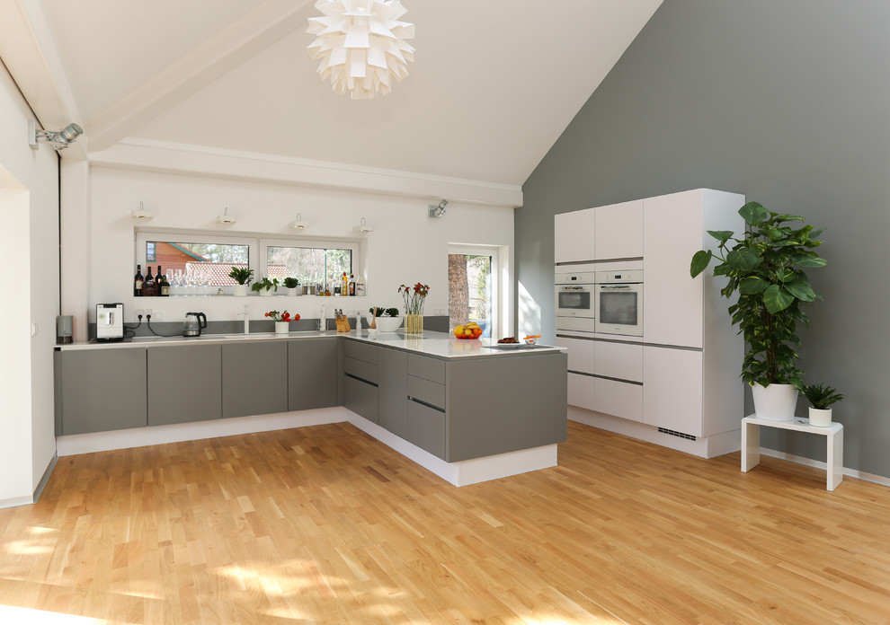Inspiration for a large contemporary grey and white l-shaped open plan kitchen in Berlin with flat-panel cabinets, grey cabinets, composite countertops, grey splashback, integrated appliances, a breakfast bar and medium hardwood flooring.