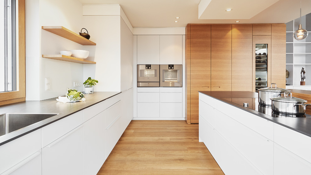 Moderne Küche in U-Form mit integriertem Waschbecken, flächenbündigen Schrankfronten, weißen Schränken, Küchenrückwand in Weiß, Küchengeräten aus Edelstahl, braunem Holzboden, Kücheninsel, braunem Boden und grauer Arbeitsplatte in Stuttgart