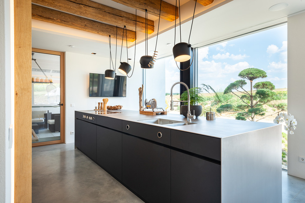 Cette image montre une grande cuisine ouverte minimaliste avec un plan de travail en inox, une crédence en bois, un électroménager noir, îlot, un sol gris et un plan de travail gris.