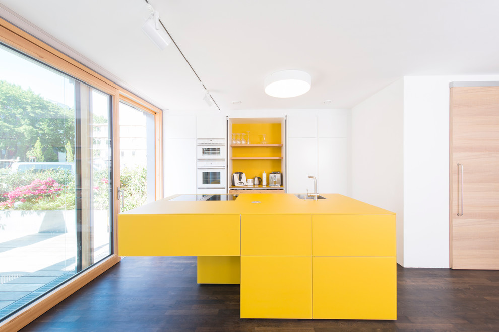Offene, Zweizeilige, Mittelgroße Moderne Küche mit flächenbündigen Schrankfronten, gelben Schränken, Küchenrückwand in Weiß, Kücheninsel, Küchengeräten aus Edelstahl, dunklem Holzboden, Waschbecken und gelber Arbeitsplatte in München