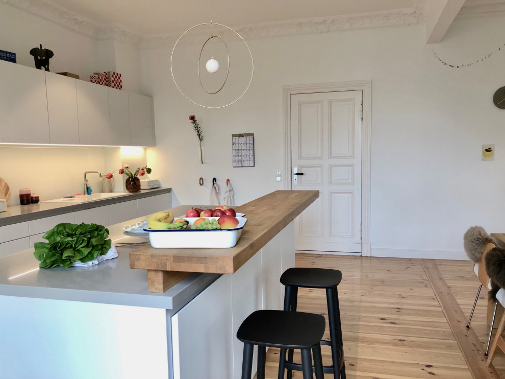 Example of a large trendy single-wall light wood floor and brown floor eat-in kitchen design in Berlin with a drop-in sink, flat-panel cabinets, white cabinets, limestone countertops, white backsplash, glass sheet backsplash, black appliances, an island and gray countertops