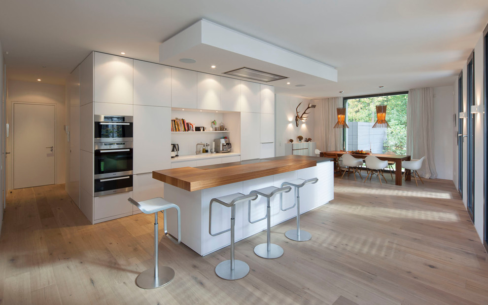Zweizeilige, Geräumige Moderne Küche mit flächenbündigen Schrankfronten, weißen Schränken, Arbeitsplatte aus Holz, Küchengeräten aus Edelstahl, braunem Holzboden und Kücheninsel in Köln