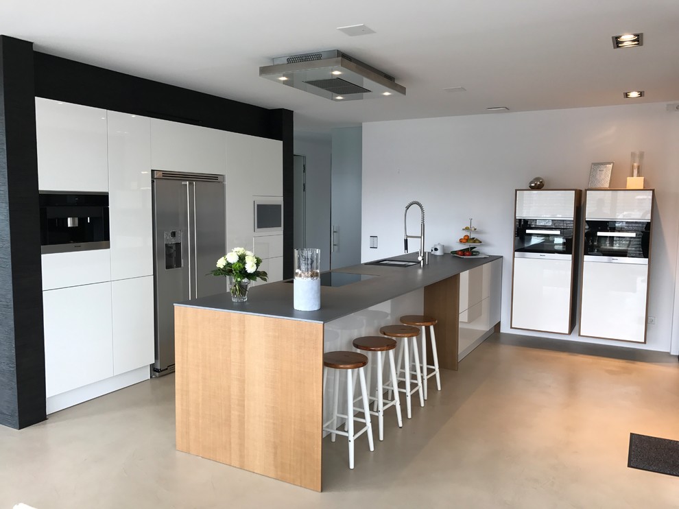Inredning av ett modernt stort grå linjärt grått kök med öppen planlösning, med en nedsänkt diskho, släta luckor, vita skåp, bänkskiva i betong, vitt stänkskydd, rostfria vitvaror, betonggolv, en köksö och beiget golv