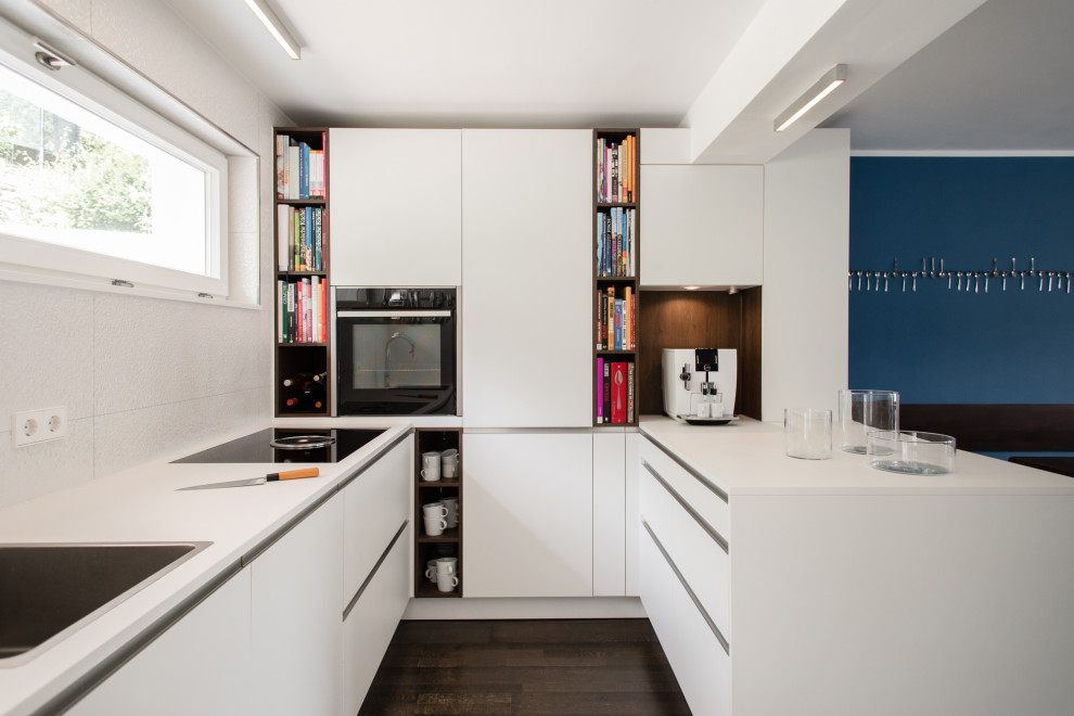 Moderne Küche in U-Form mit Einbauwaschbecken, flächenbündigen Schrankfronten, weißen Schränken, schwarzen Elektrogeräten, dunklem Holzboden, Halbinsel, braunem Boden und weißer Arbeitsplatte in Stuttgart