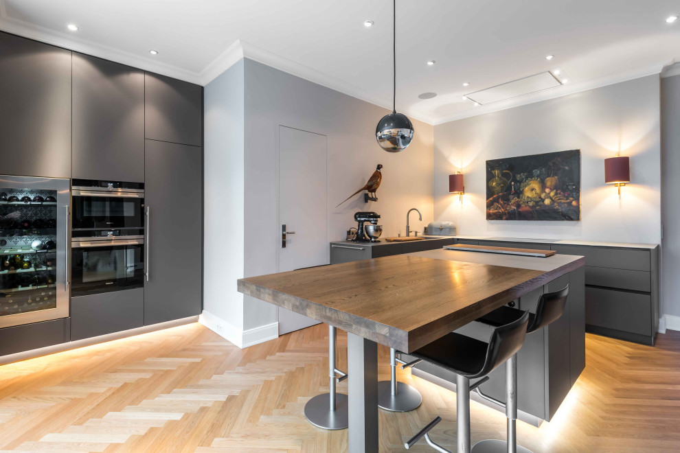 Foto på ett mellanstort funkis brun l-kök, med en undermonterad diskho, släta luckor, grå skåp, träbänkskiva, integrerade vitvaror, ljust trägolv, en köksö och beiget golv