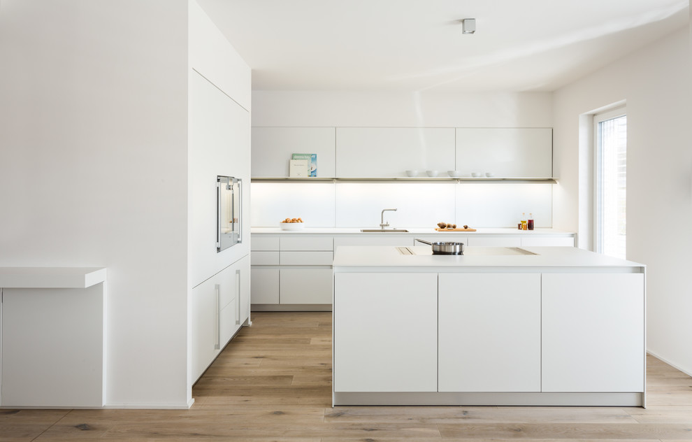 Cette photo montre une cuisine moderne en L de taille moyenne avec un placard à porte plane, des portes de placard blanches, une crédence blanche, une crédence en feuille de verre, un sol en bois brun, îlot et un sol marron.