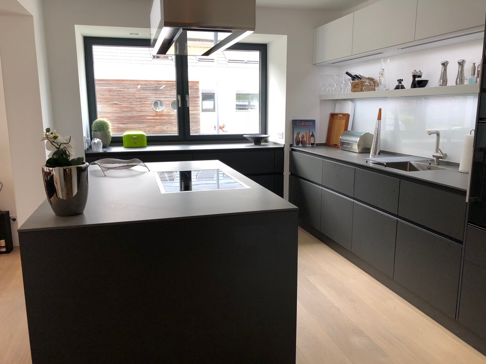 This is an example of a medium sized contemporary open plan kitchen in Other with a single-bowl sink, flat-panel cabinets, grey cabinets, concrete worktops, white splashback, glass sheet splashback, black appliances, medium hardwood flooring, an island, brown floors and grey worktops.