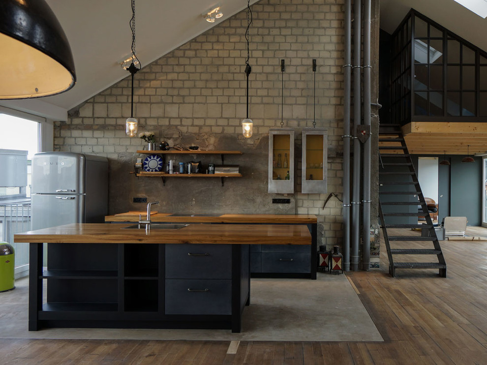 Immagine di una cucina industriale con lavello da incasso, ante nere, top in legno, paraspruzzi grigio, paraspruzzi in lastra di pietra, pavimento in legno massello medio e ante a filo