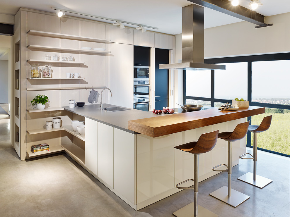 Стильный дизайн: п-образная кухня-гостиная среднего размера в современном стиле с двойной мойкой, плоскими фасадами, белыми фасадами, техникой из нержавеющей стали, бетонным полом и полуостровом - последний тренд