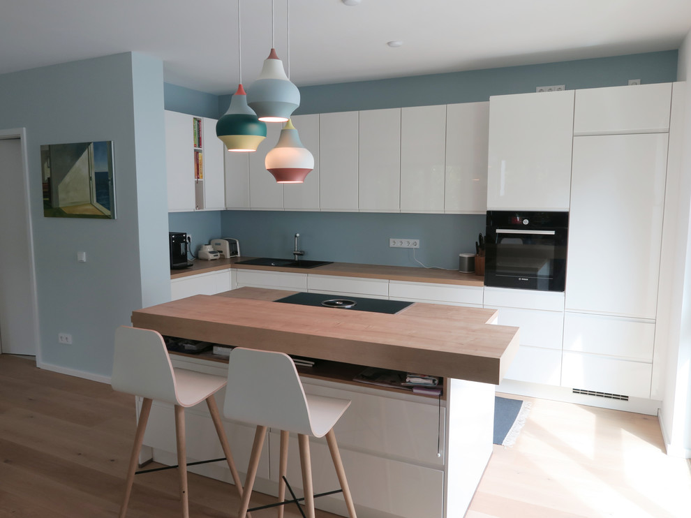 This is an example of a medium sized scandi l-shaped open plan kitchen in Cologne with a built-in sink, flat-panel cabinets, white cabinets, wood worktops, blue splashback, black appliances, light hardwood flooring, an island, beige floors and brown worktops.
