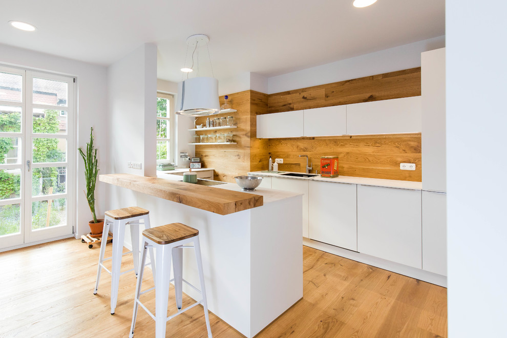 Open concept kitchen - mid-sized scandinavian u-shaped medium tone wood floor and brown floor open concept kitchen idea in Nuremberg with an integrated sink, flat-panel cabinets, white cabinets, brown backsplash, wood backsplash, a peninsula, white countertops and paneled appliances